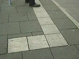 a sidewalk with an "L" shaped band of bright white tactile paving with small bars parallel to the kerb on the road