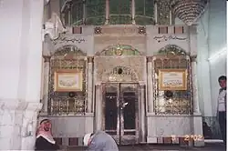 The interior of a religious building showing two men seated in front of a gated marble enclosure covered in green-tinted glass