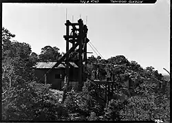 Photo of a tramway terminal located on the rim of the Grand Canyon. A wooden trestle tower about 30 or 40 feet tall with a large hut at its base can be seen; there is a large pulley visible in the tower along with several cables. Three individuals are visible to the right of the side of the tower, and appear to be in conversation with each other