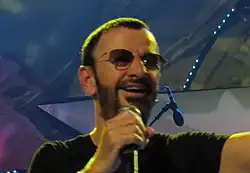 A colour photograph of Starr wearing sunglasses and singing into a microphone. The background is blue and purple.