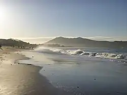 Colour photo of a long beach with foamy waves and mountains in the distance, in the morning light.