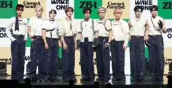Nine men standing in a row on a stage dressed in coordinated outfits featuring white tops and dark blue pants with multiple straps and buckles. Some members are wearing accessories like gloves, arm bands, and headbands.