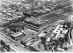 Aerial view of part of Adelaide near the Railway Station 1929. The old City Baths is in the foreground, opposite the tram on King William Road. The building between the baths and Parliament House is the Government Printing Office.