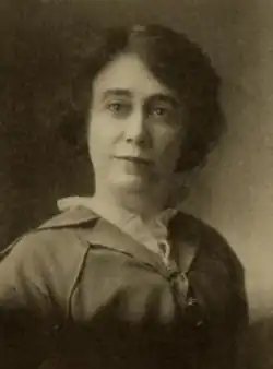 A white woman with dark hair, wearing a buttoned-up jacket over a white blouse