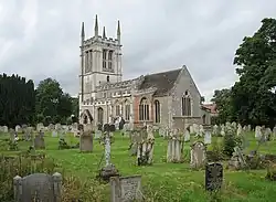 The tower of a stone church, seen from the west, at the top of which is a battlemented parapet with pinnacles. In front of the church and to the left is a lychgate