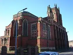 A broad church in brick with stone dressings, seen from the northeast, with a canted apse, a crocketted pinnacle, and in the distance a tower, also with crocketted pinnacles