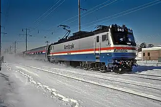 A gray electric locomotive with a black roof. At the bottom of the sides and front is a wide blue stripe, with thinner white and red stripes above.