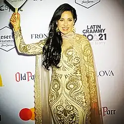 Photo of Anna Maria Mendieta at the 2021 Latin Grammy Awards After Party. She is a woman with long brown hair in a white-and-gold gown with a matching cape. She is smiling and holding a sign celebrating the Latin Grammys.
