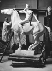 The artist places her left hand on the neck of the model of a horse whose head is above hers