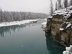 A river flows by a cliff and through a snow-covered forest.