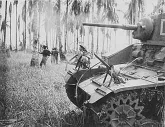 A tank and some infantry walking through knee-high grass dotted with palm trees