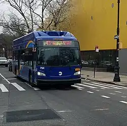 A front view of a curved mostly blue bus passing a bright yellow building