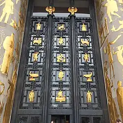 A large metal door is flanked with golden creatures and persons.
