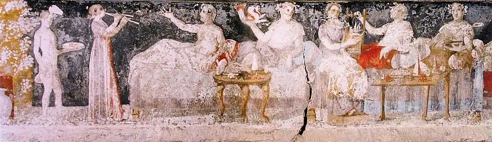 A banquet scene from a Macedonian tomb of Agios Athanasios, Thessaloniki, 4th century BC; six men are shown reclining on couches, with food arranged on nearby tables, a male servant in attendance, and female musicians providing entertainment.[111]