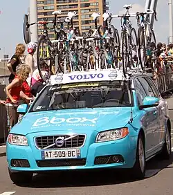 A powder blue automobile with several bicycles attached to its roof. Spectators at the roadside are looking behind the car to something out of the frame.