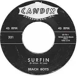 A black and white single label. The top displays the logo for the record label Candix Enterprises, Inc. The bottom says the song name "Surfin" in all caps, before listing the songwriters "B. Wilson • M. Love" and the band name "Beach Boys".