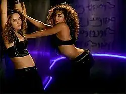 Two similarly-dressed women are dancing against a wall. From left to right, the first woman puts her hands up, while the second is putting them on the wall.