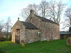 A small bare stone building seen from an angle, a lower section with a doorway at the front, and a larger, higher section with a bellcote at the back