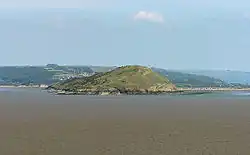 Green covered rocky land in expanse of sea. Hills behind.
