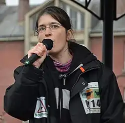 A woman with glasses holds a microphone while speaking.