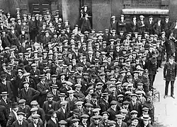 A large throng of people congregate, surrounded by police officers. Approximately half wear peaked caps; the rest wear boaters. Some smile, the rest look pensive.