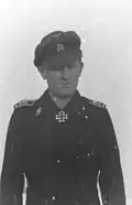 A man wearing a military uniform, peaked cap and a neck order in the shape of a cross. His cap has an emblem in shape of a human skull and crossed bones.