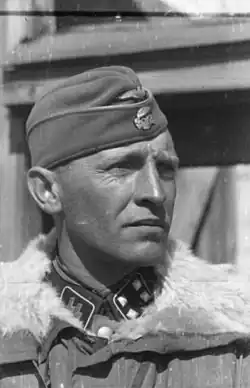 A black-and-white photograph of a man wearing a military uniform with fur collar, side cap and a neck order in shape of an Iron Cross. His cap has an emblem in shape of a human skull and crossed bones.