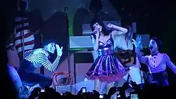 A female blond performer on stage wearing a white tuxedo and singing to a microphone in her left hand. A similarly dressed woman claps on her left. They are encircled by a group of guys in roller skates. The backdrop display disco lights in red and yellow.