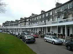 Row houses with continuous portico and stone walls on a curving street, with lines of cars parked in front