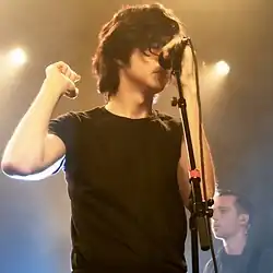 An image of the band Car Seat Headrest performing a live show at a venue called "The Showbox", with frontman Will Toledo prominently visible, and bassist Seth Dalby visible in the background. This image has been cropped into a square. Will Toledo is a white man with shaggy black hair and black frame glasses. He is holding a microphone on a stand and wearing a black t-shirt. There are stagelights in the background. This photo was taken 12 October 2018.