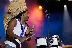Carlinhos Brown stands on the left side of the image wearing a white shirt and a headpiece, holding drum sticks in both hands. Two white drums are on the right side of the image with a microphone stand in between.