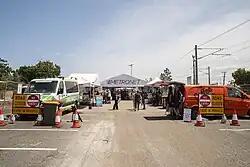 A crowd of people in the station's carpark. There are gazebos set up and a coffee van and ambulance parked.