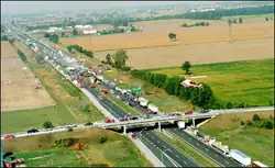 "A highway viewed from high above travels into the distance from the bottom-right to the top-left. An overpass allows a road to cross the highway near the bottom of the image. The surroundings are entirely agricultural. On the highway, several vehicles are piled into each other. The middle of the large pileup is smoking."