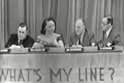 Three men and one woman sit at a long table. Behind them is a curtain. On the front of the table are the words "What's My Line?".