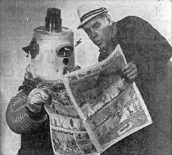 At left, a man in a robot costume made of assorted household parts with a bucket on the head, at right, a man wearing a jacket and a light-colored hat; both are holding a newspaper comics section from the Miami Herald, looking at the contents inside.