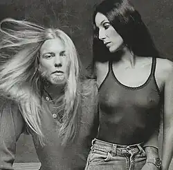 Cher, in a fitted tank top and jeans, stands next to Gregg Allman, who has long blond hair and a beard, wearing a buttoned-up shirt.