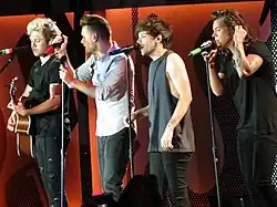 Four young adult males standing next to each other and singing into microphones as a four-piece band. From left to right: Niall Horan, Liam Payne, Louis Tomlinson, and Harry Styles.