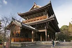 A large two-storied wooden gate with a veranda on the upper story and a hip-and-gable style roof. A small wooden building with gabled roof is placed next to it.