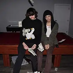 Two boys are sitting on a pool table. In front of them is money, floating in the air.