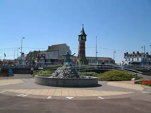A fountain with a sculpture of a person; a clock tower, people and buildings are in the background