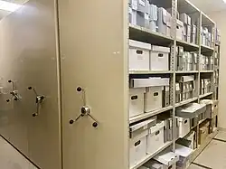 Photo of archive storage area; on the left, the hand cranks to operate shelving units; on the right, the shelves of one unit containing storage boxes