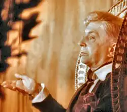 A sepia-toned photograph. A straight-faced Quentin Crisp gestures from an ornate, high-backed chair. A large, red handkerchief flops from her jacket pocket.