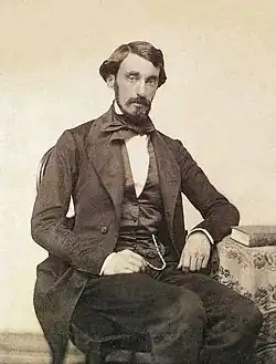 Daguerrotype portrait of a man seated next to a table and wearing black suit, vest and tie.