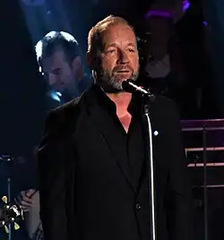 David Koller wearing a black suit, appearing to speak or sing into a microphone onstage