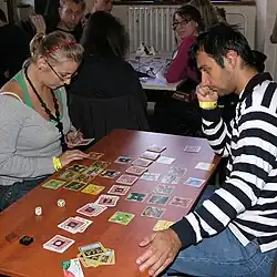 Two young people sit at a table and are in the middle of a game of the Catan Card Game