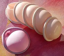 A stack of five roughly hemispherical diaphragms, each with a circular rim which is round in cross-section, all circularly symmetric. Next to them, a single diaphragm in a different colour; one portion of the near-circular rim bulges slightly, and the bowl near it has a deep, narrow moulded-in dimple, concave on the side that the main bowl is convex. The bowls are quite thin silicone rubber, a membrane that crumples under slight pressure; but the rims are more than a centimeter thick and hold their shape.