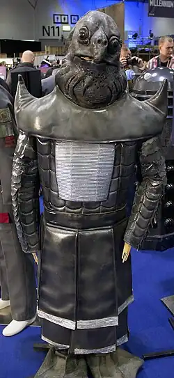 A grey, reptilian humanoid. It has a turtle-like facial structure, with black and grey armor resembling that of a samurai.