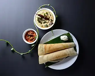 Two dosa rest next to a dollop of butter on a plantain leaf: The sauces have separate bowks.