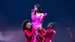 Dua Lipa performing in a pink catsuit being lifted by dancers in red outfits
