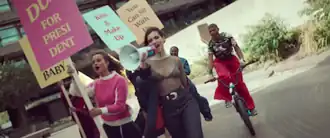 Dua Lipa singing in a megaphone with her girl group who hold rainbow flags and colourful signs with many sayings.
